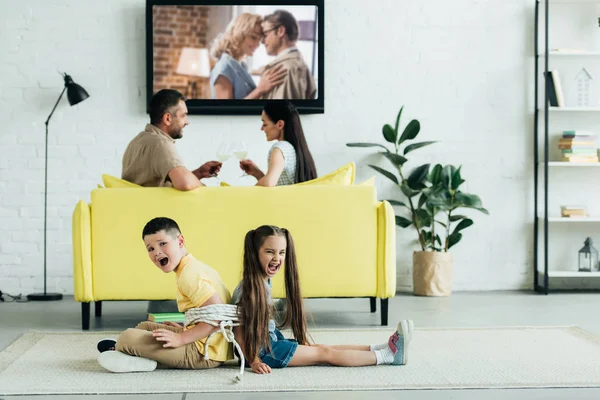 Parents resting with champagne and children screaming and sitting tied with rope on floor at home — Stock Photo