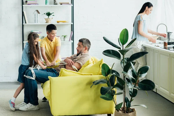 Kinder binden Vater zu Hause mit Seil, Mutter spült Geschirr — Stockfoto