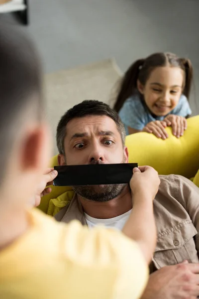 Vista aérea de los niños cerrando la boca del padre con cinta negra en casa - foto de stock