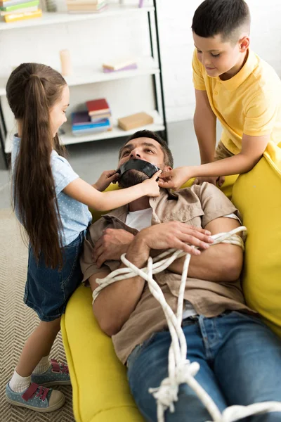 Дети закрывают рот отца черной лентой дома — стоковое фото