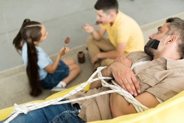 Geschwister essen Schokolade und Vater liegt gefesselt auf Sofa zu Hause, Elternschaft Konzept — Stockfoto