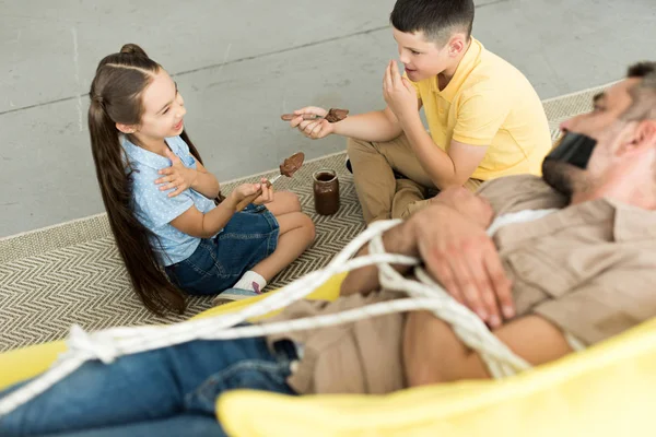 High angle view of children eating chocolate and father lying tied on sofa at home — Stock Photo