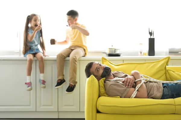 Дети едят шоколад и отец лежит привязанный на диване дома — стоковое фото