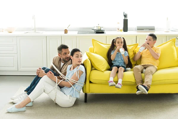 Pais amarrados sentados perto do sofá e crianças comendo sobremesa em casa — Fotografia de Stock