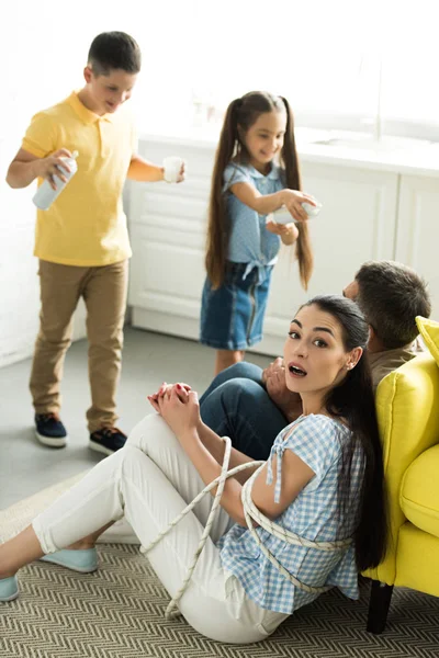 Tied parents sitting near sofa and kids scaring them with cream at home — Stock Photo