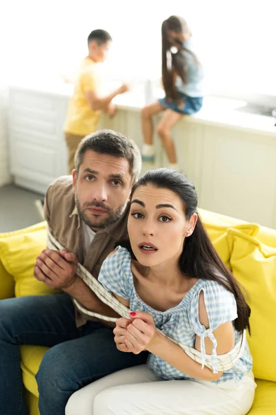 Scared tied parents sitting on sofa and looking at camera at home, parenthood concept — Stock Photo
