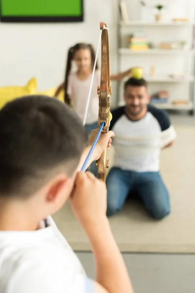 Enfoque selectivo de los niños jugando con el padre atado y fingiendo disparar con arco de juguete en casa - foto de stock