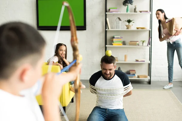 Crianças brincando com pai amarrado e fingindo tiro com arco de brinquedo na sala de estar — Fotografia de Stock