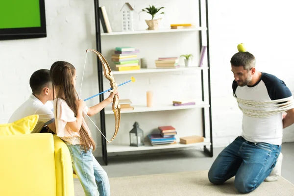 Visão lateral de crianças brincando com pai amarrado e fingindo atirar com arco de brinquedo em casa — Fotografia de Stock