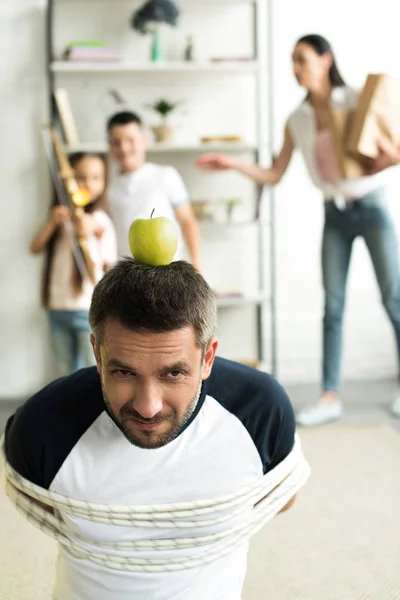 Tied father sitting on floor with apple on head for target at home, parenthood concept — Stock Photo