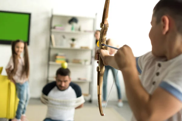 Siblings playing with tied father and pretending shooting with toy bow at home — Stock Photo