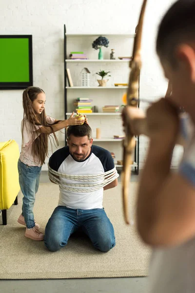 Enfants désobéissants jouant avec le père attaché et faisant semblant de tirer avec l'arc de jouet à la maison — Photo de stock