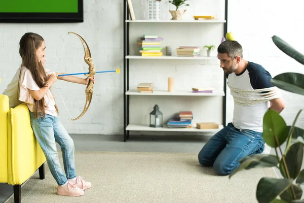 Side view of daughter playing with tied father and pretending shooting with toy bow at home — Stock Photo