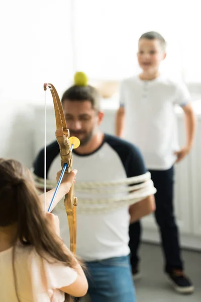 Foco seletivo de crianças brincando com pai amarrado e fingindo atirar com arco de brinquedo em casa — Fotografia de Stock