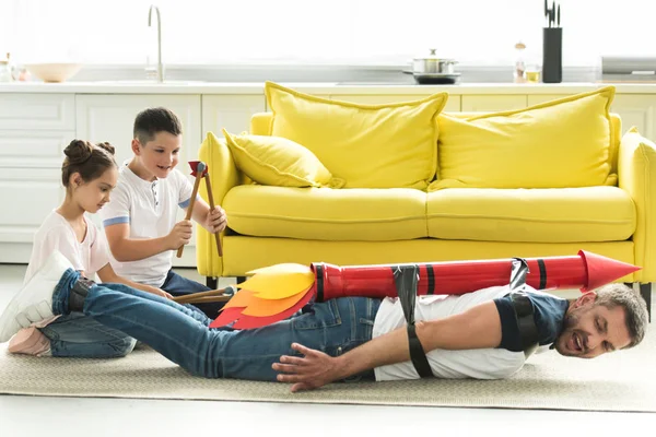 Tied father with rocket toy lying on floor and disobedient children playing with him — Stock Photo