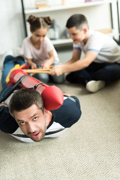 Tied daddy with rocket toy lying on floor and children playing with him — Stock Photo