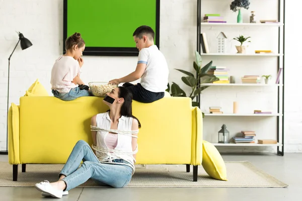 Side view of children eating popcorn and mother sitting tied with rope on floor at home — Stock Photo