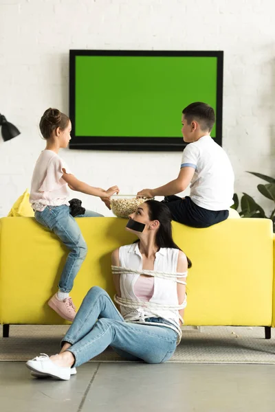 Children eating popcorn and mother sitting tied with rope on floor at home — Stock Photo