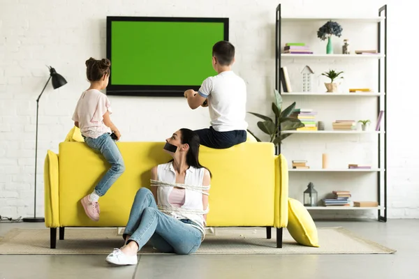 Children playing video game and mother sitting tied with rope on floor at home — Stock Photo