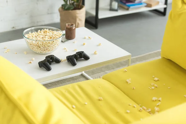 Скляна чаша з попкорном і джойстиками на столі, жовтий диван у вітальні — стокове фото