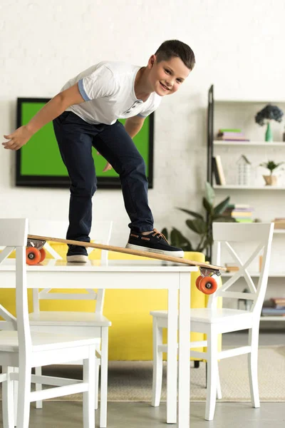 Vorpubertärer Junge steht zu Hause auf Skateboard auf Tisch und blickt in Kamera — Stockfoto