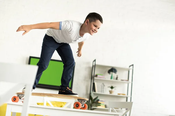 Smiling pre-adolescent boy standing on skateboard on table at home — Stock Photo