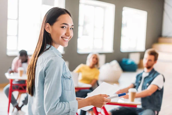 Young asian student girl with paper standing in front of classmates and looking at camera — Stock Photo