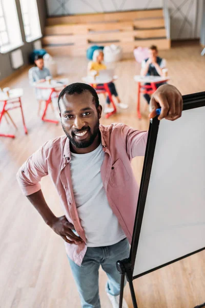 Jeune professeur afro-américain appuyé sur le tableau blanc à la salle de conférence avec des étudiants flous assis sur le fond — Photo de stock