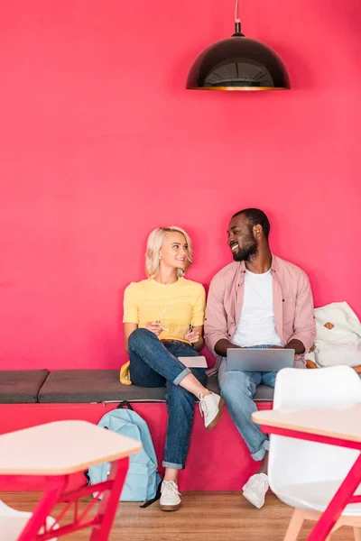 Multiethnische junge Studenten arbeiten mit Laptop, während sie unter roter Wand sitzen — Stockfoto