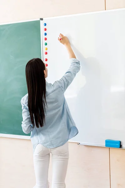 Vista trasera de asiático estudiante chica escritura en whiteboard durante lección - foto de stock