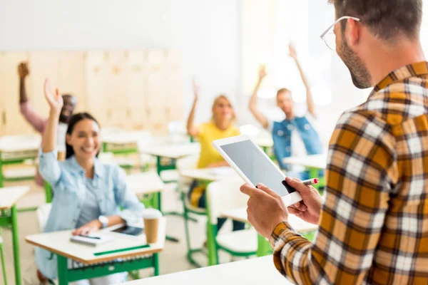 Group of young students raising up hands to answer on teachers question while he standing with tablet on foreground — Stock Photo