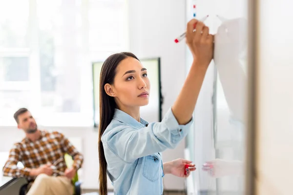 Young asian student girl writing on whiteboard during lesson with blurred teacher on background — Stock Photo