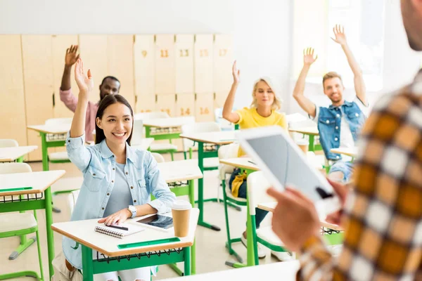 Молоді студенти піднімають руки, щоб відповісти на питання вчителів, стоячи з планшетом на передньому плані — стокове фото