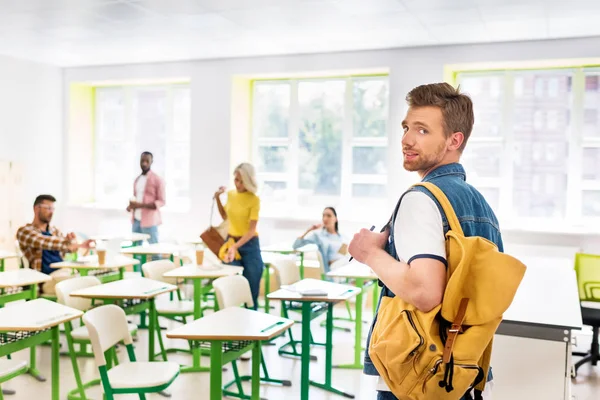 Стильный молодой студент в классе колледжа с размытыми одноклассниками на заднем плане — стоковое фото