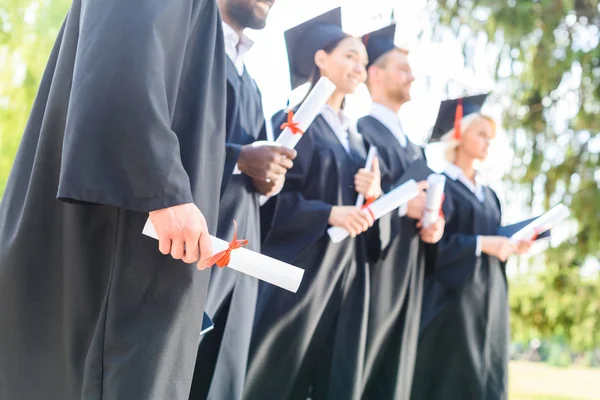 Tiro recortado de los estudiantes graduados en capas y sombreros que poseen diplomas laminados - foto de stock