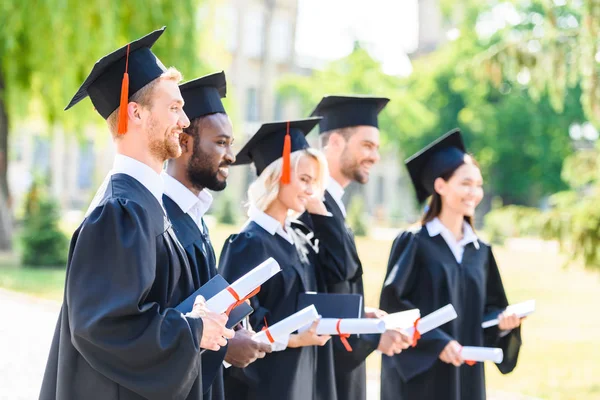 Glückliche Absolventen in Umhängen und Hüten mit gerollten Diplomen — Stockfoto