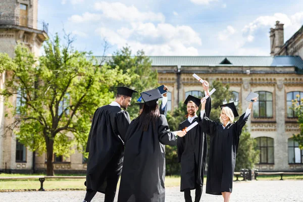 Jóvenes estudiantes graduados con diplomas saludándose en el jardín universitario - foto de stock
