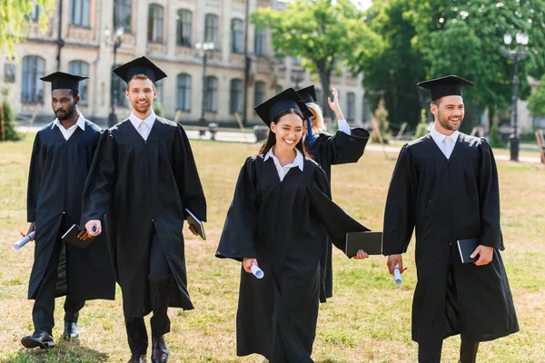 Jóvenes estudiantes graduados en capas caminando por el jardín universitario - foto de stock