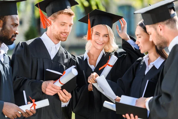 Estudiantes graduados multiétnicos sonrientes en capas con diplomas - foto de stock