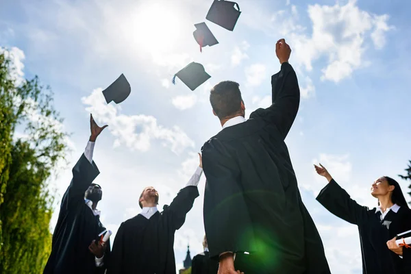 Bottom view of young graduated students throwing up hats in front of blue sky — Stock Photo