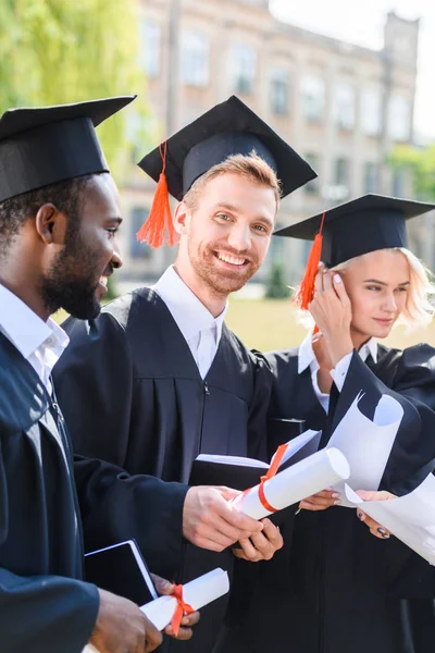 Estudantes multiétnicos felizes graduados em capes com diplomas — Fotografia de Stock