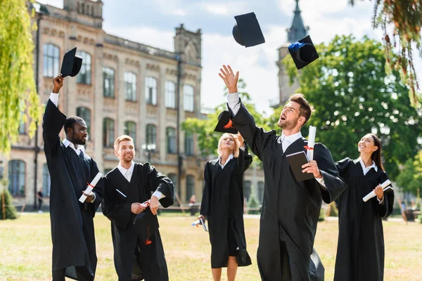 Jóvenes felices estudiantes graduados vomitando gorras de graduación en el jardín universitario - foto de stock