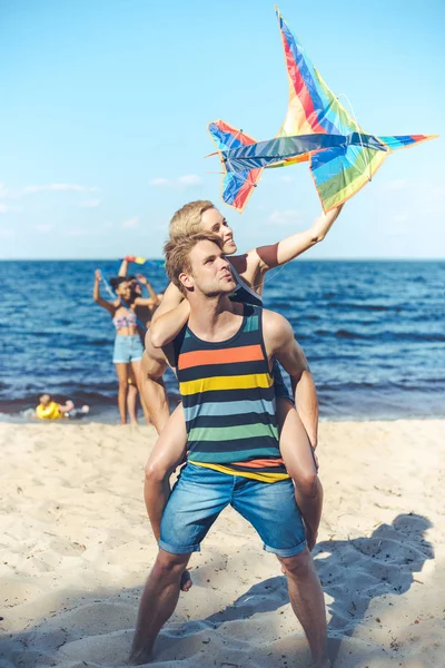 Селективный фокус пары с красочным воздушным змеем с многорасовыми друзьями позади проводить время на песчаном пляже — стоковое фото