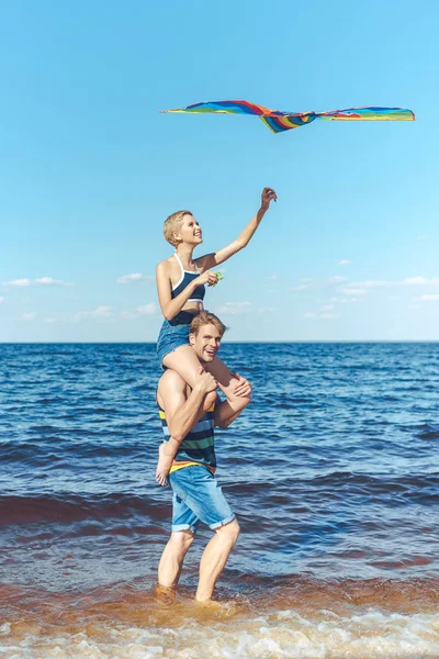 Young woman with colorful kite sitting on boyfriends shoulders on beach — Stock Photo