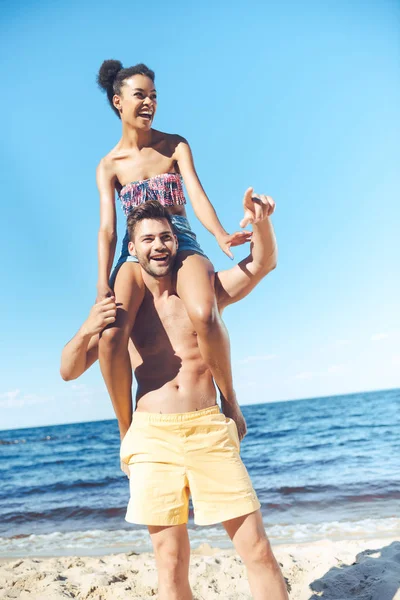 Cheerful african american woman sitting on boyfriends shoulders on beach — Stock Photo