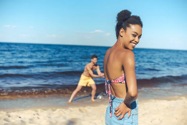 Focalizzazione selettiva di donna afro-americana sorridente e fidanzato caucasico dietro sulla spiaggia di sabbia — Foto stock