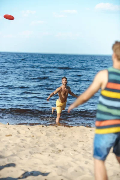 Selective focus of young men playing with flying disc on sandy beach — Stock Photo