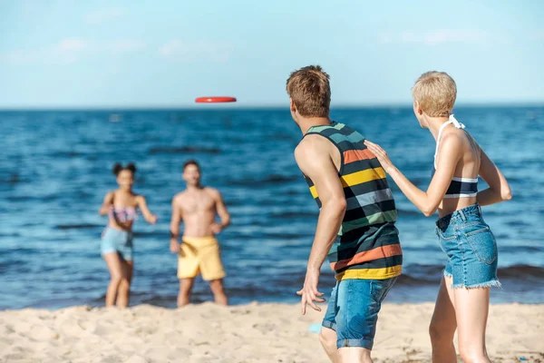 Foco seletivo de amigos multirraciais brincando com disco voador juntos na praia — Fotografia de Stock