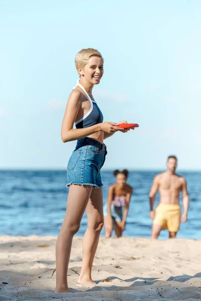 Selective focus of interracial friends playing with flying disk on sandy beach — Stock Photo