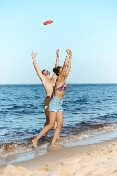 Young multicultural couple playing with flying disc on beach on summer day — Stock Photo